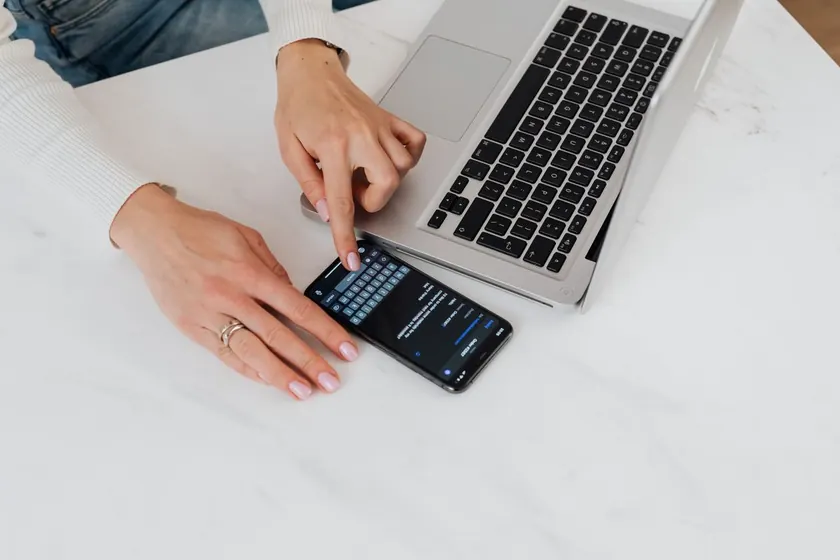 Manos usando un teléfono móvil junto a un portátil sobre una mesa blanca