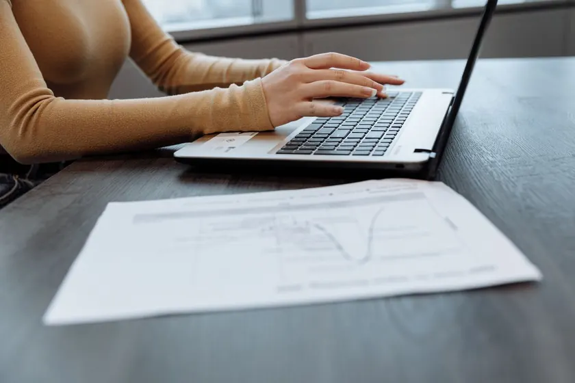 Manos tecleando en un portátil con un documento impreso sobre la mesa